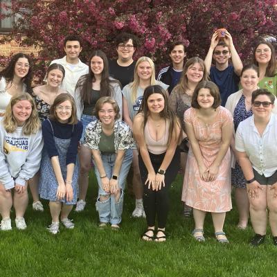  The image shows a group of fifteen young adults posing outdoors on a grassy area, with a flowering tree and brick building in the background. They are smiling and dressed casually. One person in the back is holding a small orange object above their head.